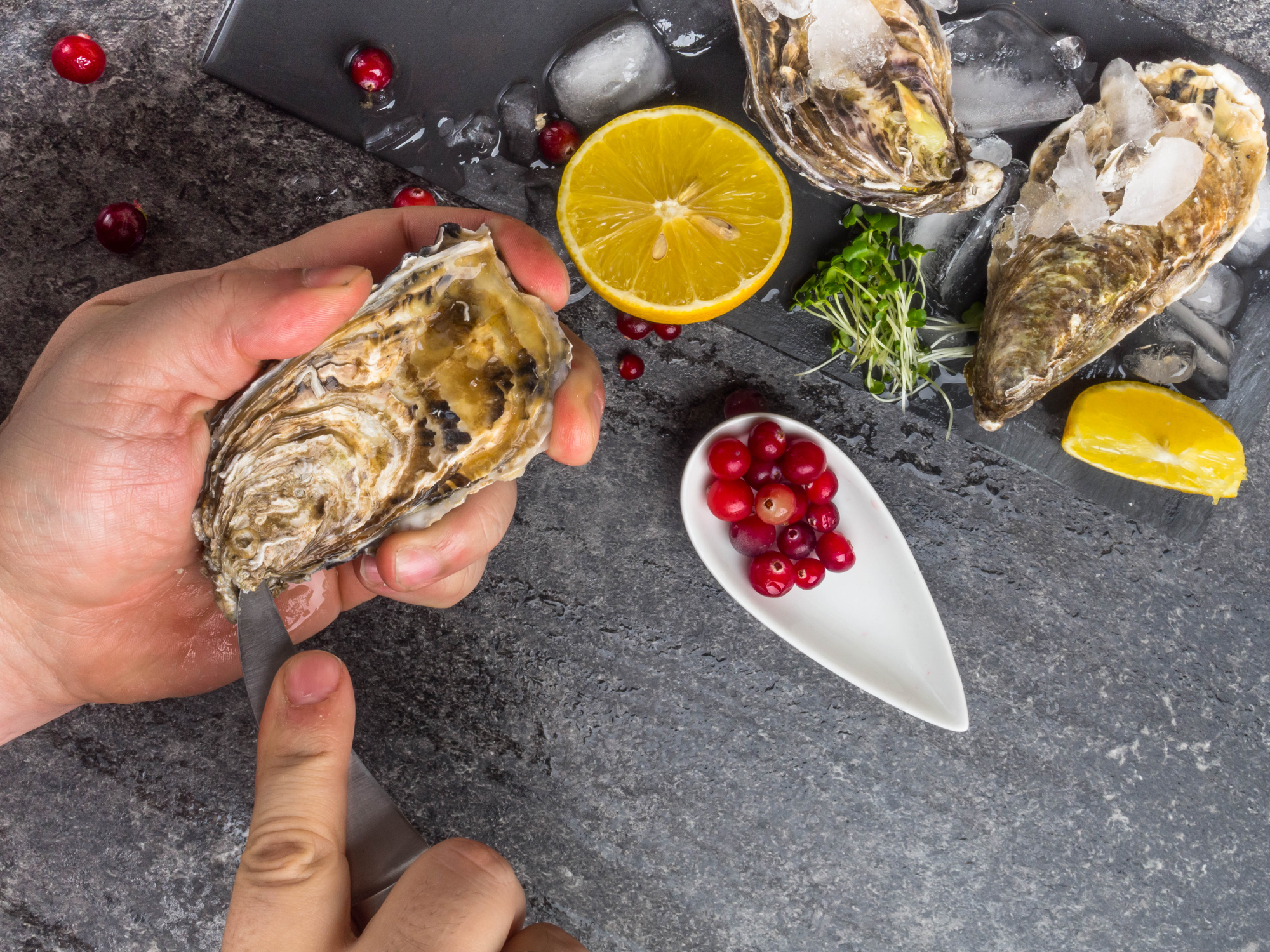 A man with a knife opens a fresh oyster. Dark stone background with lemon, ice, berries, selective focus. A man with a knife opens a fresh oyster. Dark stone background with lemon, ice, berries, selective focus. Food for men health, rich in protein, iodine, zinc